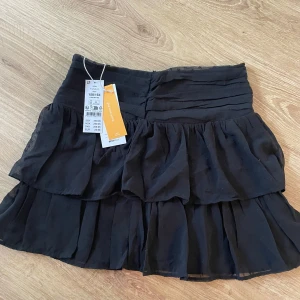 Svart volangkjol från Gina Tricot - Supersöt svart kortkjol från Gina Tricot med flera lager volanger och lätt rynkade detaljer upptill. Kjolen har en luftig känsla och är perfekt för dig som gillar en trendig och lekfull stil. Passar till allt och är enkel att matcha med olika toppar.