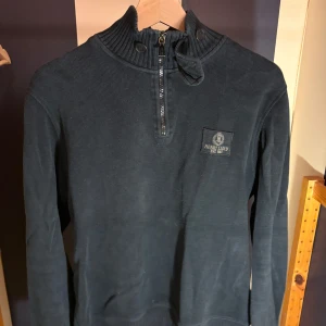 Mörkblå half zip tröja Henri Lloyd - Snygg mörkblå tröja från Henri Lloyd med ribbad krage och half zip framtill. Tröjan har ett broderat märke på bröstet och diskreta ränder på insidan av kragen. Långärmad och tillverkad i mjuk bomull, perfekt för kyliga dagar.
