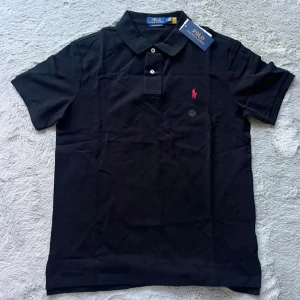 Svart Polo Ralph Lauren piké M - Svart pikétröja från Polo Ralph Lauren i modellen Custom Slim Fit. Klassisk krage, tre knappar och röd broderad logga på bröstet. Tillverkad i mjuk bomull och perfekt för en clean och stilren look.