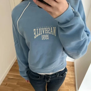 Blå sweatshirt från Bershka - Ljusblå sweatshirt från Bershka med vit text framtill. Tröjan har vita detaljer längs ärmarna och ribbade muddar vid hals, ärmslut och nederkant. Skön och avslappnad passform, perfekt till jeans. Liten i storleken- passar bäst XS. 