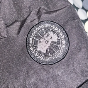 Svart dunjacka från Canada Goose XL - Säljer en svart dunjacka från Canada Goose i storlek XL. Jackan har en klassisk pufferstil med huva och Canada Goose-logga på ärmen. Perfekt för kalla dagar med sin tjocka vaddering och snygga, stil, saknar en knapp där fram och den man hänger upp den på är lite trasig