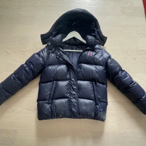 Mörkblå pufferjacka från Polo Ralph Lauren - Säljer en mörkblå pufferjacka från Polo Ralph Lauren med avtagbar huva och röd broderad logga på bröstet. Jackan har dragkedja framtill och är vadderad för extra värme. Perfekt för kalla dagar och riktigt snygg streetstil.                                Jackan är köpt från kidsbrandstore. Det är en barnmodell, storleken passar för 12-14 åringar. Jackan har andvända i två vinter säsonger. Som man kan se på sista bilden har den lite defekter runt armarna.