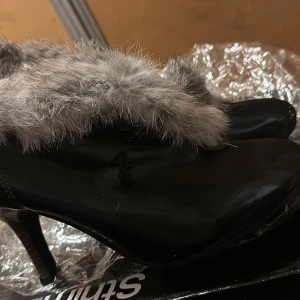 Svarta stövletter med fuskpäls - Snygga svarta stövletter med hög klack och mjuk grå fuskpäls runt skaftet. Skorna har en elegant siluett med spetsig tå och är perfekta för dig som vill sticka ut. Materialet är blankt och ger en lyxig känsla.