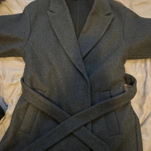 Grå kappa med bälte från Vero Moda - Snygg grå kappa från Vero Moda i klassisk modell med bälte i midjan och breda slag. Kappan har två fickor framtill och är tillverkad i ett mjukt ullblandat material. Perfekt för dig som vill ha en stilren och tidlös look under höst och vinter.