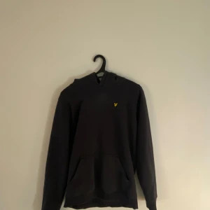 Svart hoodie från Lyle & Scott - Svart hoodie från Lyle & Scott med klassisk gul logga på bröstet. Nypris 749 kr. Tröjan har huva, ficka och långa ärmar. Perfekt för chill dagar eller när du vill ha en enkel och stilren look. Hoodien är för 12-13 åringar. Passar för folk som är allt från 160cm till 170 cm lång. Varning! hoodien har en liten defekt som man ser på en av bilderna 