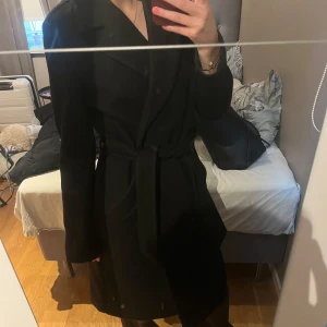 Svart kappa med bälte - Original pris omkring 800kr. Säljer en svart kappa med klassisk krage och knytbälte i midjan. Jackan är lång och har en stilren, tidlös look som passar perfekt till lager-på-lager. Långärmad och i en rak modell som ger en snygg siluett.