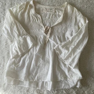 Vit blus från Odd Molly - Supersöt vit blus från Odd Molly med broderade detaljer och smock upptill. Blusen har trekvartsärmar med virkade kanter och ett snyggt knyt vid halsen. Luftig och flowy passform, perfekt för ljusa dagar. Hittar tyvärr inte storlek men skulle säga att den passar som en M.