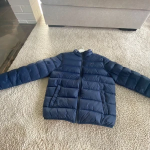Mörkblå dunjacka från Tommy Hilfiger - Snygg mörkblå dunjacka från Tommy Hilfiger med diskret logga på bröstet och dragkedja framtill. Jackan har quiltad design, hög krage med logotyp och röd insida. Perfekt för kalla dagar när du vill hålla stilen och värmen. Köpt för 1500kr på kidsbrandstore