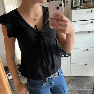 Svart blus med knyt framtill - Säljer en svart blus med v-ringning och knyt framtill. Blusen har korta ärmar med lätt volang och är tillverkad i ett mjukt tyg som faller snyggt. Perfekt att styla med jeans eller kjol för en trendig look.