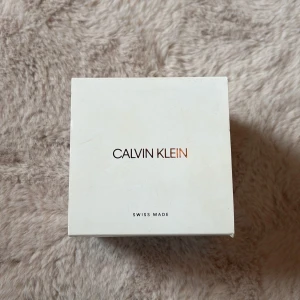 Calvin Klein silverfärgad meshklocka - Säljer en stilren Calvin Klein klocka med silverfärgad urtavla och svart mesharmband. Urtavlan har minimalistisk design med CK-logga och svarta visare. Klockan levereras i original ask. Perfekt accessoar för dig som gillar enkel och modern stil.