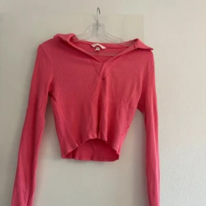 Rosa ribbad croppad topp med krage - Säljer en rosa croppad topp från H&M med lång ärm och snygg krage. Toppen är ribbad och har en v-ringad öppning framtill. Perfekt för dig som gillar trendiga och bekväma plagg med lite edge.