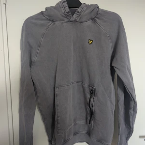 Grå hoodie från Lyle & Scott  - Säljer en grå hoodie från Lyle & Scott med klassisk huva och känguruficka med dragkedja. Tröjan har lång ärm och den ikoniska gula loggan på bröstet. Strl motsvarar 164/170 (xs i herr)