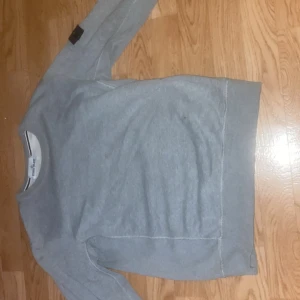 Grå Stone Island hoodie - Snygg grå sweatshirt från Stone Island med klassisk patch på ärmen. Tröjan har rund halsringning, långa ärmar och är tillverkad i mjuk bomull. Perfekt för en clean och stilren look, passar till jeans eller joggers.