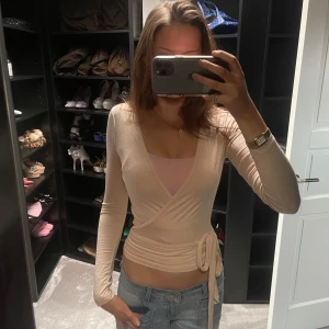 Beige omlottopp - Snygg beige omlottopp med långa ärmar och knytning i sidan. Perfekt för en stilren look. Liknande från excere, köptes på nån TikTok sida som hade liknande excere! Knappt använd men är väldigt snygg 🥹💕