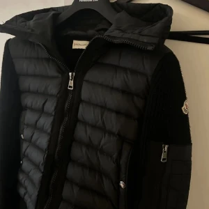 Svart cardigan från Moncler - Svart cardigan från Moncler med stickade ärmar och huva. Jackan har quiltad framsida, dragkedja och Moncler-logga på ärmen. Snygg kombination av syntet och stickat material, perfekt för dig som gillar exklusiv streetstyle.