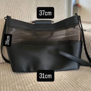 Svart axelväska med transparent detalj - Stilren svart axelväska med cool transparent panel upptill och rundade handtag. Väskan har både axelrem och innerpåse i tyg med snörning och dragkedja. Perfekt storlek för allt du behöver ha med dig och ger en modern vibe.