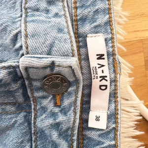 Blå jeans från NA-KD, storlek 38 - Säljer ett par ljusblå jeans från NA-KD i klassisk femficksmodell med bruna detaljer och tydliga sömmar. Jeansen har rak passform och är tillverkade i jeansmaterial med snygg tvättad look. Perfekt för dig som gillar en chill och stilren vibe.