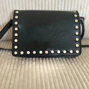 Svart axelväska med nitar från Don Donna - Snygg svart axelväska från Don Donna i skinnimitation med silverfärgade nitar runt locket. Väskan har justerbar axelrem, magnetlås och ett huvudfack med innerficka som stängs med dragkedja. Perfekt storlek för det viktigaste och ger en cool vibe till din outfit.