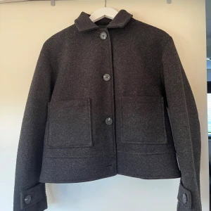 Mörkgrå  kappa från 157, stl S - Säljer en stilren mörkgrå kappa från 157. Jackan har raka linjer, stora knappar framtill, två rejäla fickor och klassisk krage. Perfekt för dig som gillar minimalistisk och clean look. Fodrad insida för extra komfort.