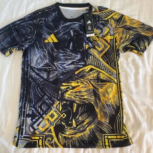 Real Madrid Special fotbollströja - Snygg Real Madrid fotbollströja från Adidas med ett galet grafiskt mönster i svart, gul och grå. Tryckt lejonmotiv och broderad klubbmärke på bröstet. Tröjan är kortärmad och har rund hals. Tillverkad i lätt, ventilerande Aeroready-material.
