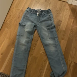 Blå cargo jogger från Jack, stl 164 - Snygga ljusblå cargo jogger-byxor från Jack med stora fickor på sidorna och dragsko i midjan. Byxorna har en avslappnad passform och är gjorda i mjukt bomullsmaterial. Perfekta för en chill och street-inspirerad look.