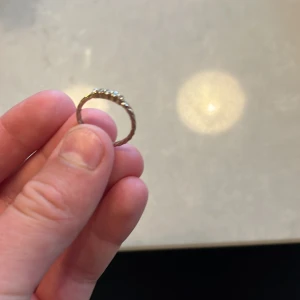 Tunn guldtonad ring med stenar - Säljer en tunn ring i guldton med tvistad design och små glittrande stenar på ovansidan. Ringen har en enkel look som passar perfekt för dig som gillar stilrena smycken med lite extra bling.
