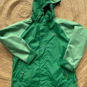 Grön regnjacka från Trekmates - Snygg tvåfärgad grön regnjacka från Trekmates med löstagbar huva och dragkedja framtill. Jackan har långa ärmar, justerbara muddar med kardborre och två fickor med dragkedja. Perfekt för blöta dagar och outdoor-äventyr.