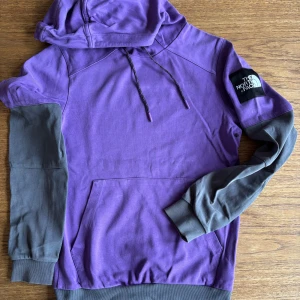 Lila hoodie från The North Face XS - Snygg lila hoodie från The North Face med grå kontrastärmar och muddar. Tröjan har huva med snörning, stor magficka och logga på ärmen. Perfekt för dig som gillar streetwear och vill sticka ut med färg.