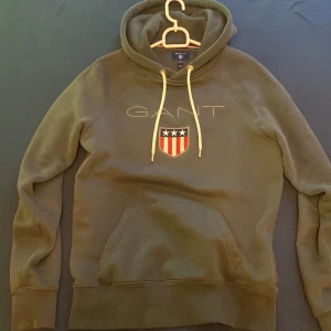 Mörkgrön hoodie från GANT XS - Säljer en mörkgrön hoodie från GANT i storlek XS. Tröjan har broderad GANT-logga och ett sköldmärke framtill, vita snören i huvan och klassisk känguruficka. Perfekt för dig som gillar stilren och sportig look.