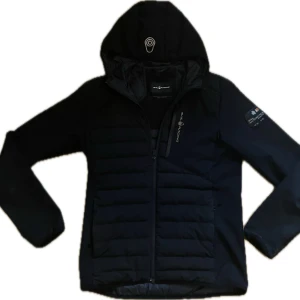 Svart Sail Racing pufferjacka med huva - Svart pufferjacka från Sail Racing med huva och snygga detaljer. Jackan har quiltade partier, dragkedja framtill och Sail Antarctica Expedition-märke på ärmen. Perfekt för kalla dagar och har en sportig vibe. Fodrad insida och diskret logga på bröstet.