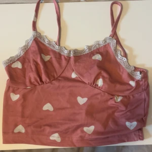 Rosa linnetopp med hjärtan och spets - Supersöt rosa linnetopp med vita hjärtan och tunna axelband. Toppen har en fin spetskant i ljusgrått längs urringningen och är croppad i modellen. Perfekt för dig som gillar gulliga detaljer och en romantisk vibe.