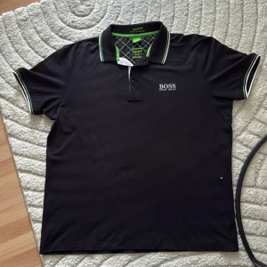 Svart Hugo Boss pikétröja - Svart pikétröja från Hugo Boss med vit och grön detalj på krage och ärmslut. Klassisk polokrage med knappar och broderad logga på bröstet. Tillverkad i polyester för en sportig känsla och fukttransport. Perfekt för en clean och stilren look.