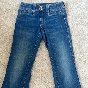 Blå bootcut jeans med avtagbara ben - Säljer ett par blå jeans med bootcut passform och avtagbara ben, så du kan enkelt göra om dem till shorts. Jeansen har klassisk femficksdesign, dubbla knappar i midjan och snygga slitningar. Perfekta för dig som vill ha en unik och flexibel stil.