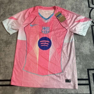 Barcelona rosa fotbollströja - Snygg Barcelona fotbollströja från Nike i rosa och vitt med tunna linjer och mönster. Tröjan har korta ärmar, V-ringad krage med blå detalj och tryckt Spotify-logga på bröstet. Materialet är lätt och andas, perfekt för träning eller match.