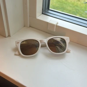 Vita solglasögon med bruna glas - Snygga solglasögon med vit, lite tjockare båge och bruna glas. Modellen har en klassisk, något fyrkantig form och små silvriga detaljer vid hörnen. Perfekt för att ge din look en fräsch och modern touch under soliga dagar.