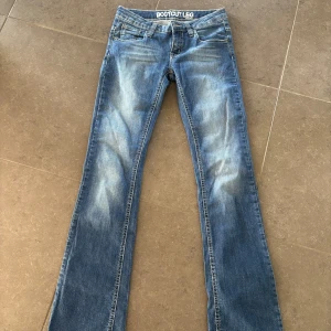 Blå bootcut jeans låg midja - Snygga blå jeans med bootcut-ben och låg midja. Jeansen har klassisk femficksdesign, ljusa slitningar framtill och markerade sömmar. 
