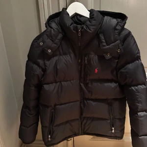 Svart pufferjacka Polo Ralph Lauren - Svart pufferjacka från Polo Ralph Lauren med huva, dragkedjor på fickor och bröstficka samt ikonisk röd logga på bröstet. Jackan är vadderad och har en sportig vibe, perfekt för kalla dagar. Snygg och enkel att matcha med allt.