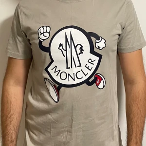 Beige Moncler t-shirt - Snygg beige t-shirt från Moncler med en stor, tecknad logga framtill. T-shirten är kortärmad och har en klassisk passform. Tillverkad i 100% bomull för en skön känsla. Inget kvitto medgår köpet och därför kan jag tänka mig gå ner någon hundralapp i pris! 