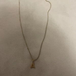 Guldigt halsband med bokstav A från Pilgrim - Snyggt halsband i guldton från Pilgrim med ett hängsmycke i form av bokstaven A. Kedjan är tunn och elegant, perfekt för att ge en personlig touch till din stil. Märkt med Pilgrim vid låset för extra detalj.