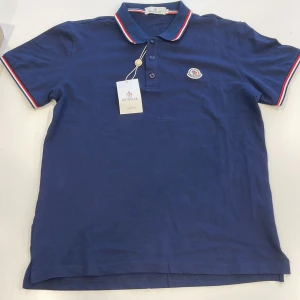 Blå Moncler piké t-shirt - Snygg marinblå piké från Moncler med klassisk krage och korta ärmar. T-shirten har vita och röda ränder på krage och ärmslut, samt Moncler-logga broderad på bröstet. Tillverkad i mjuk bomull och har knappar framtill. Passar s 