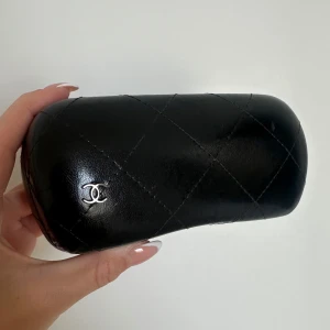 Svart glasögonfodral - Snyggt svart glasögonfodral i quiltat skinn med silverfärgad logga på framsidan. Fodralet har en stilren och lyxig look och passar perfekt för att skydda dina glasögon.