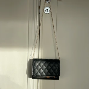 Svart axelväska med kedja - Snygg svart axelväska med quiltat mönster och guldfärgad kedja. Väskan har en stilren design med metall-detalj framtill och är perfekt för att lyfta vilken outfit som helst.