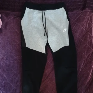 Nike mjukisbyxor i svart och grå - Snygga mjukisbyxor från Nike med svart och ljusgrå färgblock, klassisk logga på benet och snörning i midjan. Perfekta för chill eller träning, med skön passform och mjukt material. Elastiska muddar vid benslut och praktiska fickor på sidorna.