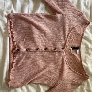 Rosa kofta från H&M Divided XS - Supersöt ljusrosa kofta från H&M Divided i storlek XS. Koftan är ribbstickad med långa ärmar, knappar framtill och volangkant nertill. Perfekt att slänga på sig över ett linne eller t-shirt för en chill look.