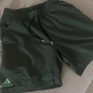 Mörkgröna Adidas shorts med snörning - Snygga mörkgröna shorts från Adidas med elastisk midja och svart snörning med gröna detaljer. Perfekta för träning eller chill, med klassisk Adidas-logga på benet. Lätta och sköna i polyester som andas.