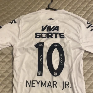 Santos FC Neymar Jr. matchtröja - Säljer en vit Santos FC fotbollströja från Umbro med Neymar Jr. och nummer 10 på ryggen. Tröjan har tryckta sponsorer, klubbmärke med stjärnor och klassisk rund hals. Materialet är lätt och andas, perfekt för dig som vill ha en ikonisk look på planen.