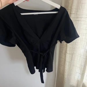 Svart omlottopp från Gina Tricot - Svart omlottopp från Gina Tricot med v-ringning och knyt i midjan. Toppen har korta ärmar och en figurnära passform som framhäver midjan. Perfekt att styla med jeans eller kjol för en trendig look.