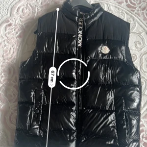 Svart dunväst från Moncler - Snygg svart dunväst från Moncler med glansig finish och broderad logga på bröstet. Västen har hög krage, dragkedja framtill med Moncler-tryck och två fickor med knappar. Perfekt för lager-på-lager och streetstyle.