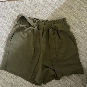 Olivgröna shorts med knyt i midjan - Olivgröna shorts med hög midja och avtagbart knytband i midjan. Inga knappar eller gylf men resår. 