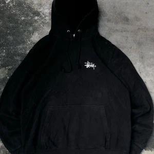 Svart Stüssy hoodie med stor logga - Svart hoodie från Stüssy i storlek L med fet vit logga på ryggen och mindre logga på bröstet. Klassisk känguruficka, huva med snörning och skön passform. Tillverkad i 80% bomull och 20% polyester – perfekt för streetwear-stilen.